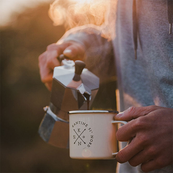 Camping-Tasse mit Kaffee, beiger Emaillebecher, Hand am Henkel