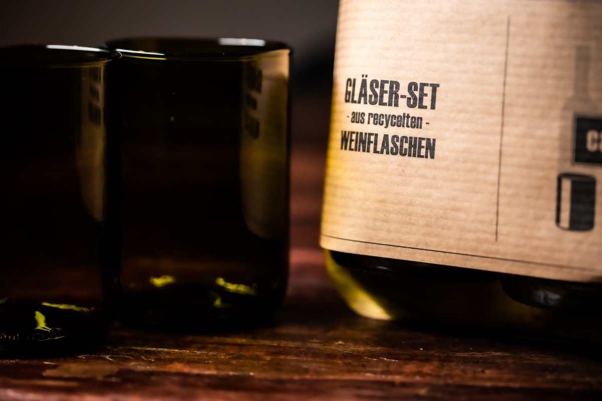 Trinkglas-Set im Karton, daneben ein dunkler Glasbecher mit breitem Rand